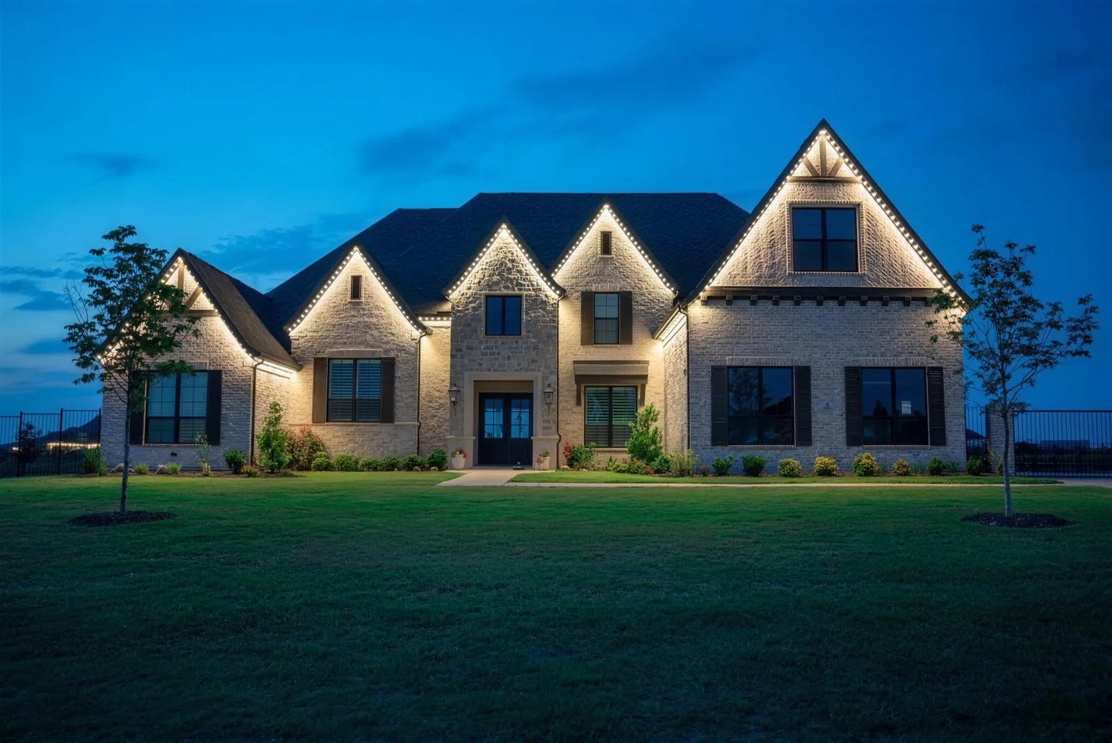 The same brick home at blue hour with warm white permanent lighting along every gable